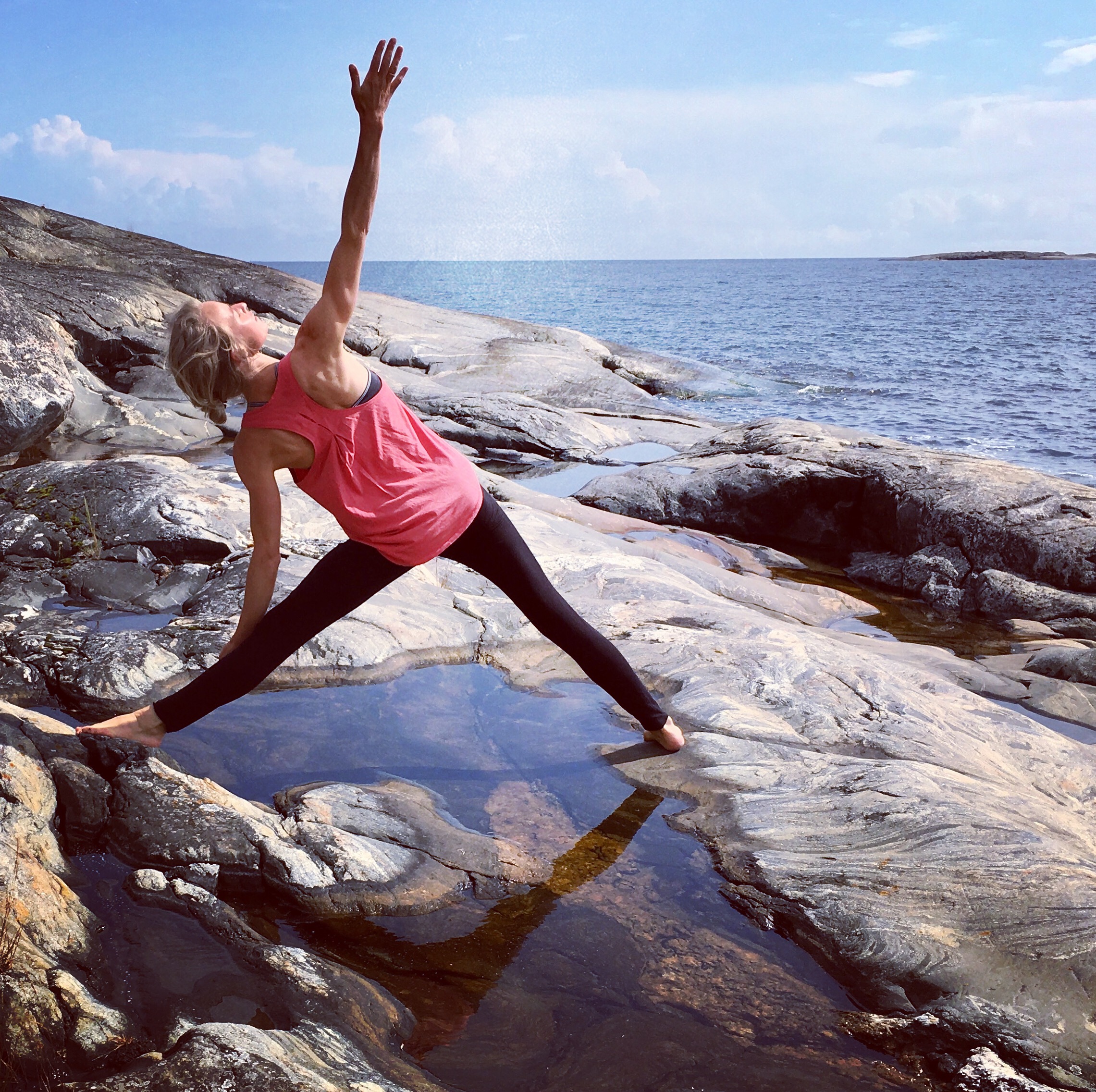 Hanna in warrior pose on sea rocks, blue sky and water behind her